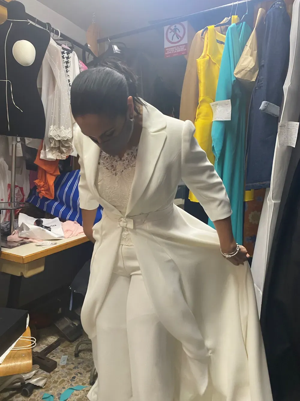 Mujer con traje de novia blanco se ajusta la falda en una tienda de ropa, rodeada de estantes de ropa.
