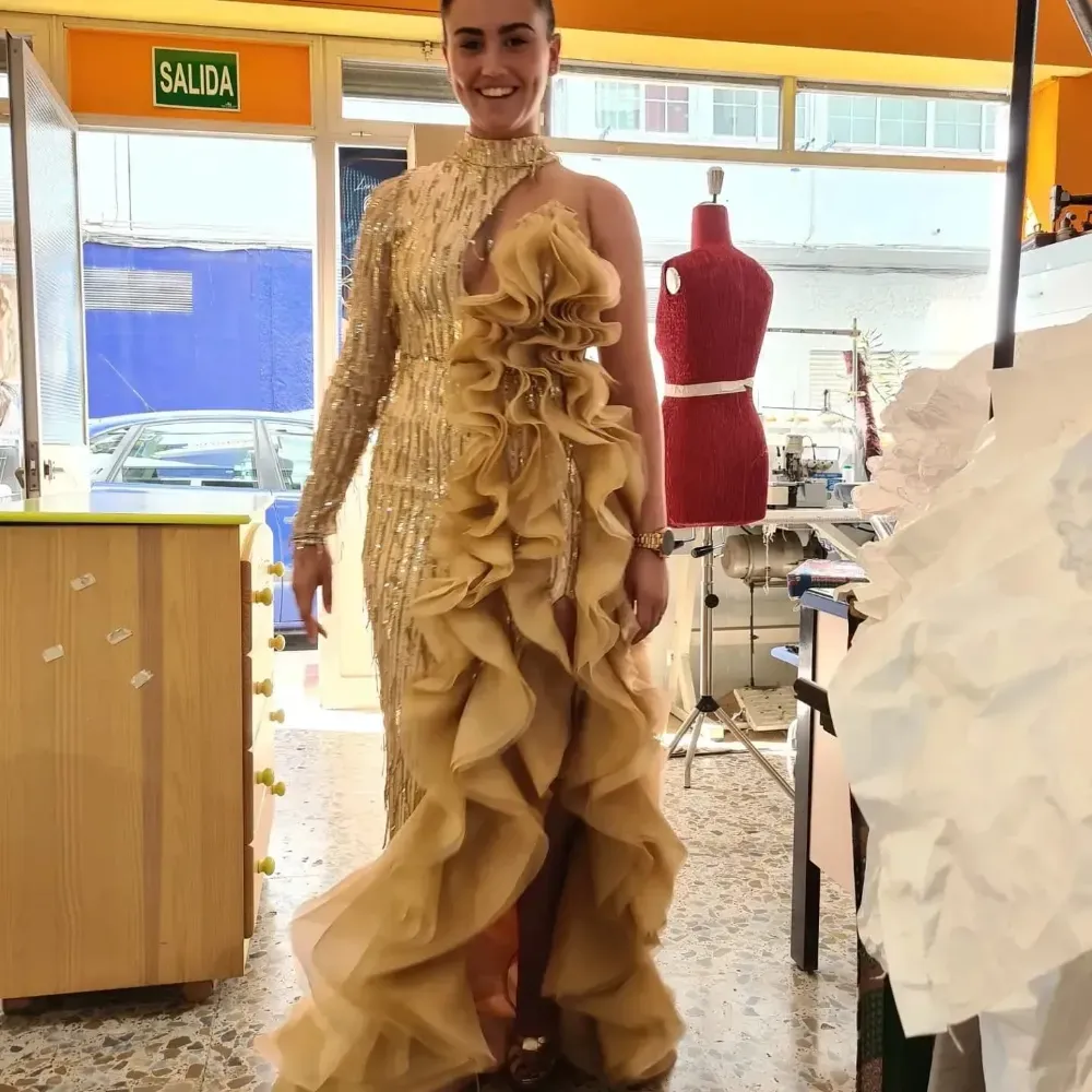 Mujer con vestido dorado posa en un taller. Vestido de un solo hombro, con volantes y lentejuelas. Sonriendo.
