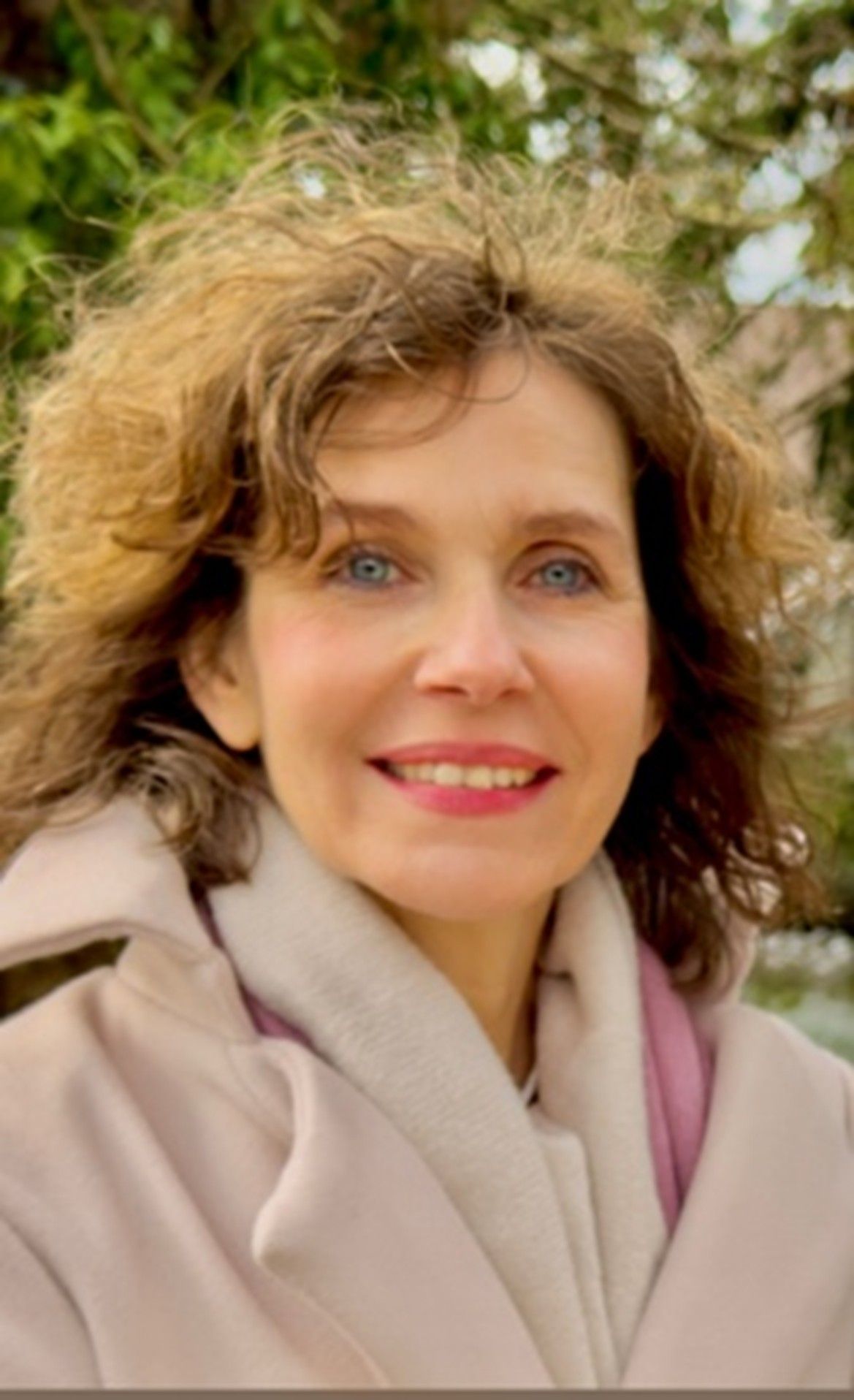 Frau mit kurzem, lockigem braunem Haar lächelt, trägt ein weißes Hemd, eine Perlenkette und rote Ohrringe.