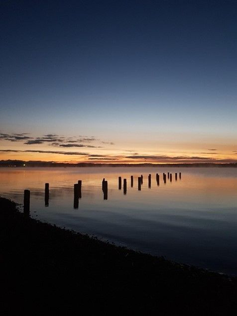Sonnenaufgang über dem Wasser mit Holzpfosten, orangefarbenem und blauem Himmel, Küstensilhouette.