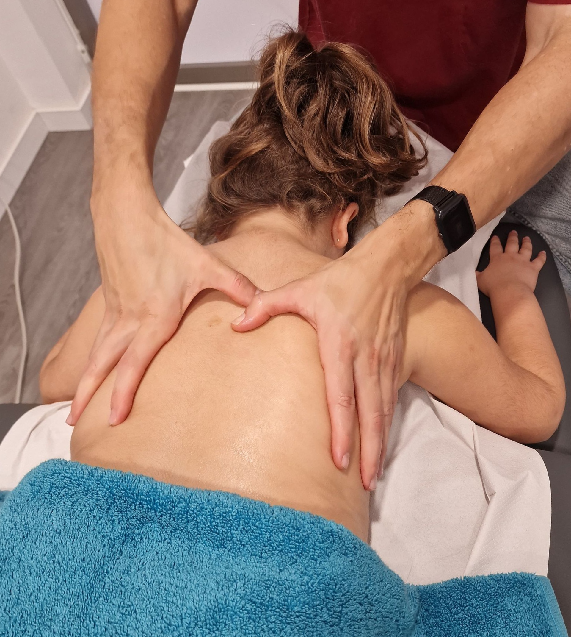 Persona recibiendo un masaje de espalda de un terapeuta. Tiene las manos en la espalda. Se ven una toalla azul y una camisa roja.