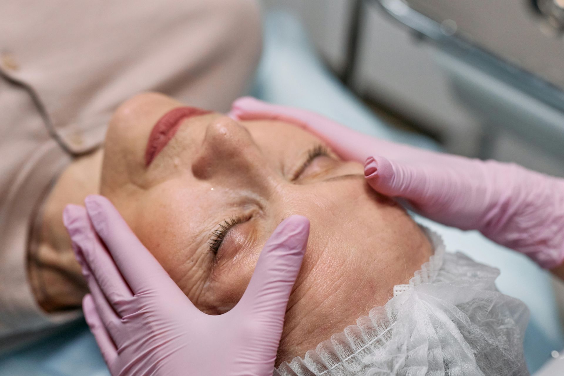 Mujer recibiendo un tratamiento facial, con las manos enguantadas de color rosa sobre su cara, en un salón.