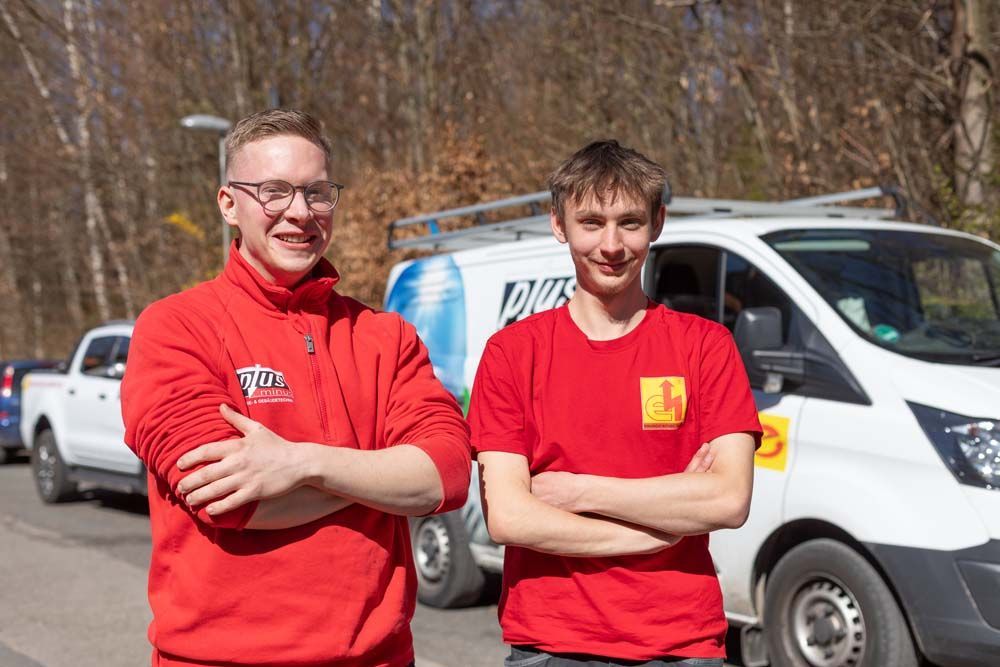 Zwei Männer in roten Hemden stehen vor einem Lieferwagen. Einer trägt eine Brille, beide haben die Arme verschränkt. Im Freien.