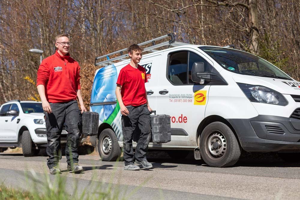An einem sonnigen Tag kommen zwei Männer in roten Hemden und mit Werkzeug aus einem Lieferwagen, der am Straßenrand geparkt ist.