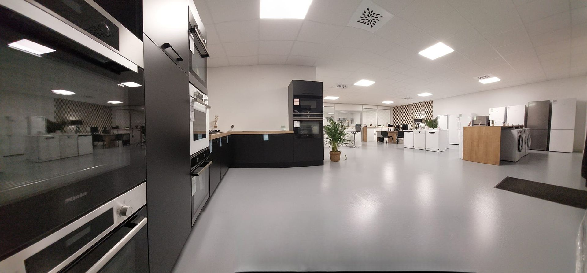 Une salle d'exposition présentant des appareils de cuisine, notamment des fours, des réfrigérateurs et des lave-linge. Le sol est gris.