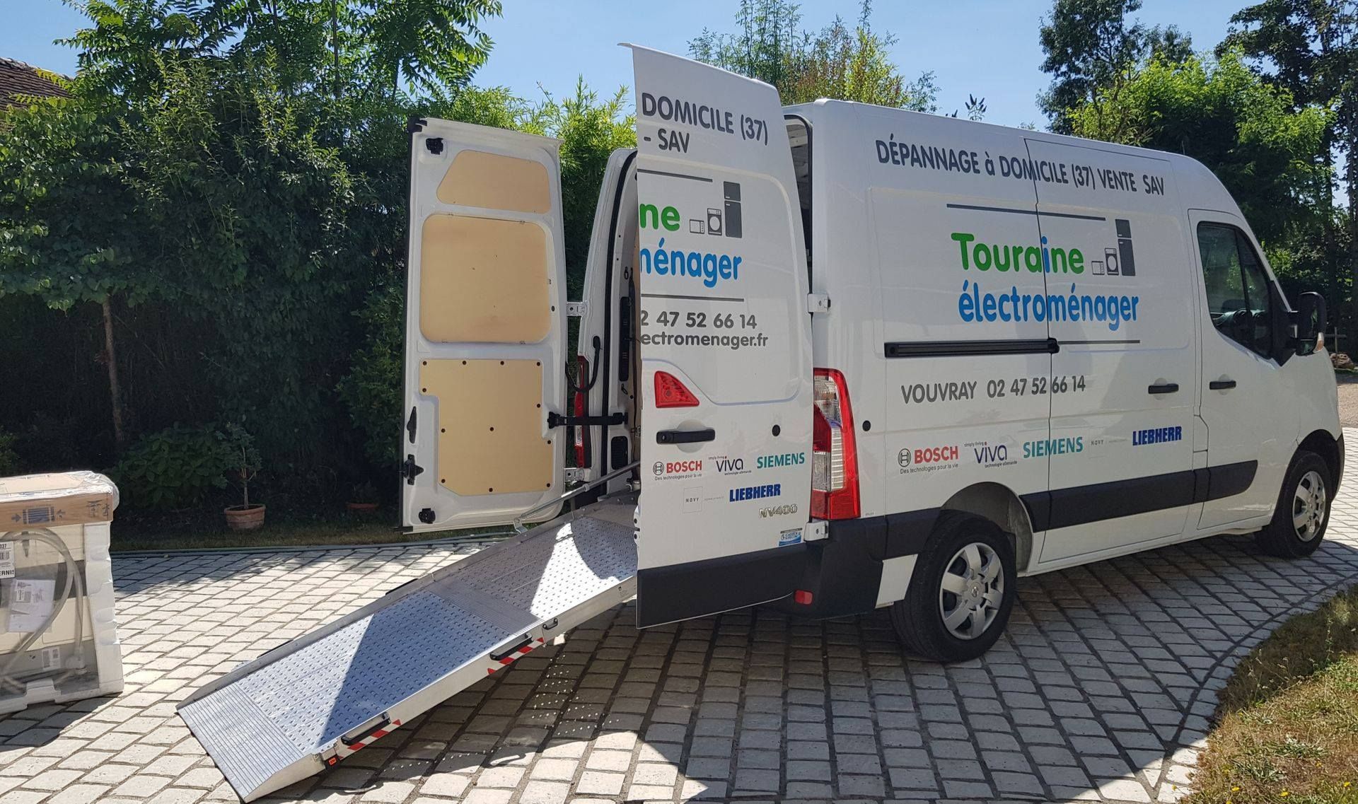 Fourgonnette de service blanche avec rampe arrière ouverte, stationnée sur une allée pavée devant une cour.