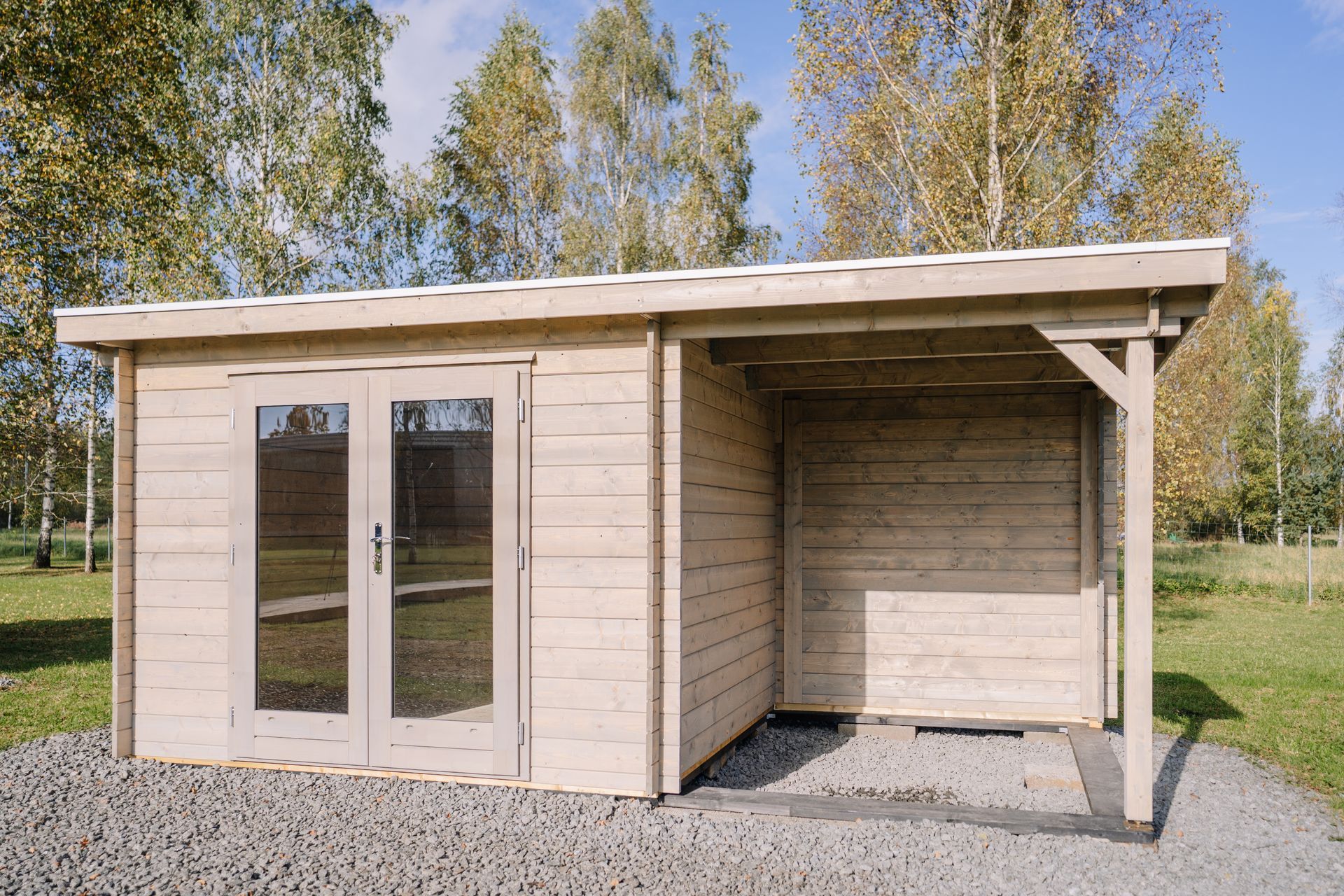 Abri de jardin en bois avec double porte vitrée et porche couvert attenant, terrain en gravier gris, entouré d'arbres.
