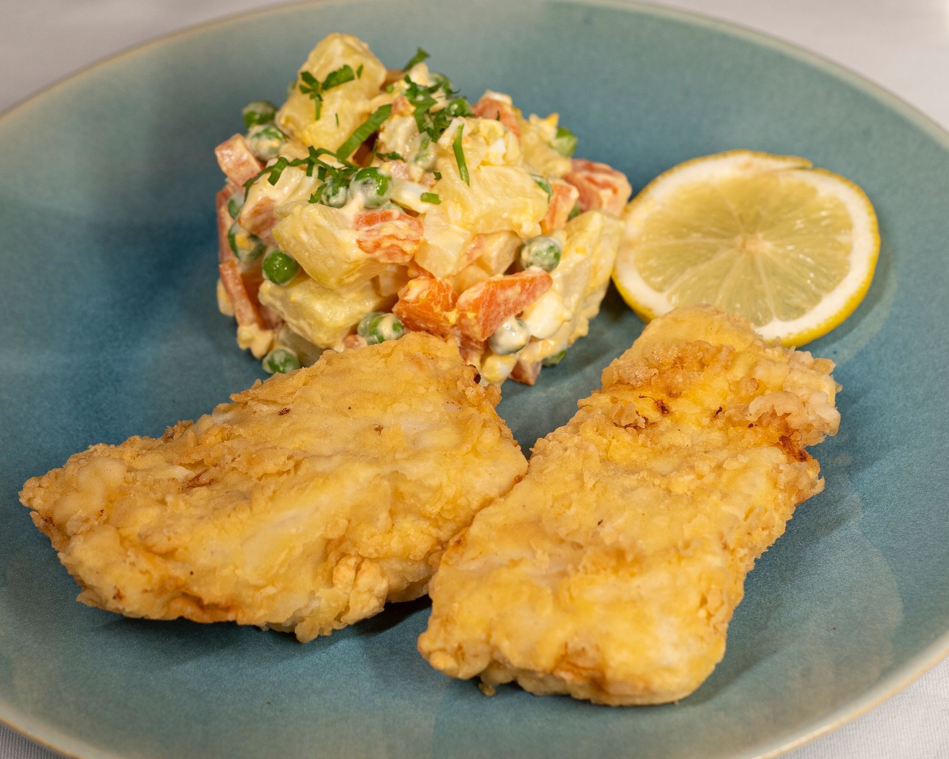 Um prato azul coberto com peixe frito, salada de batata e uma rodela de limão.