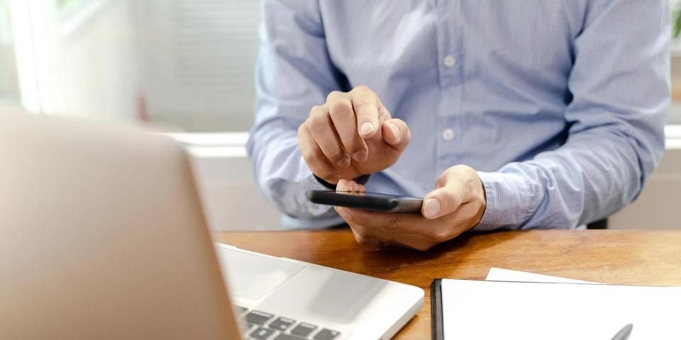 Evelin Buck Steuerberatung, Männerhand tippt auf Smartphone, Laptop am Tisch