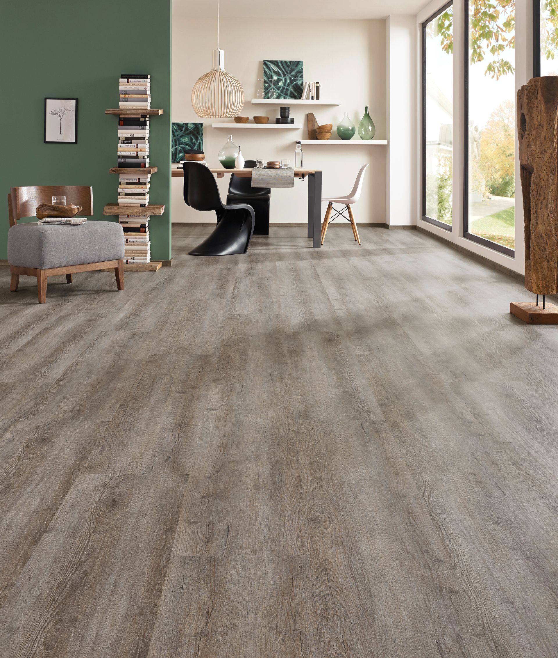 Geräumiges Zimmer mit grauem Holzoptikboden, Essbereich und großen Fenstern. Grüne Akzentwand und Dekorationsgegenstände.