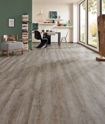 Geräumiges Zimmer mit grauem Holzoptikboden, Essbereich und großen Fenstern. Grüne Akzentwand und Dekorationsgegenstände.