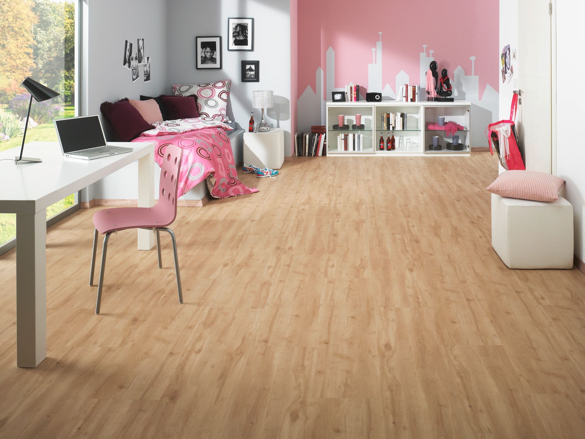 Ein Schlafzimmer mit hellem Holzboden, einer rosa Wand, einem weißen Schreibtisch und einem Bett mit einer gemusterten Bettdecke.