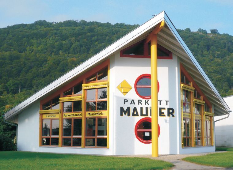 Weißes Gebäude mit roten und gelben Akzenten, Schild „Parkett Maurer“. Grüner Rasen, Bäume im Hintergrund.