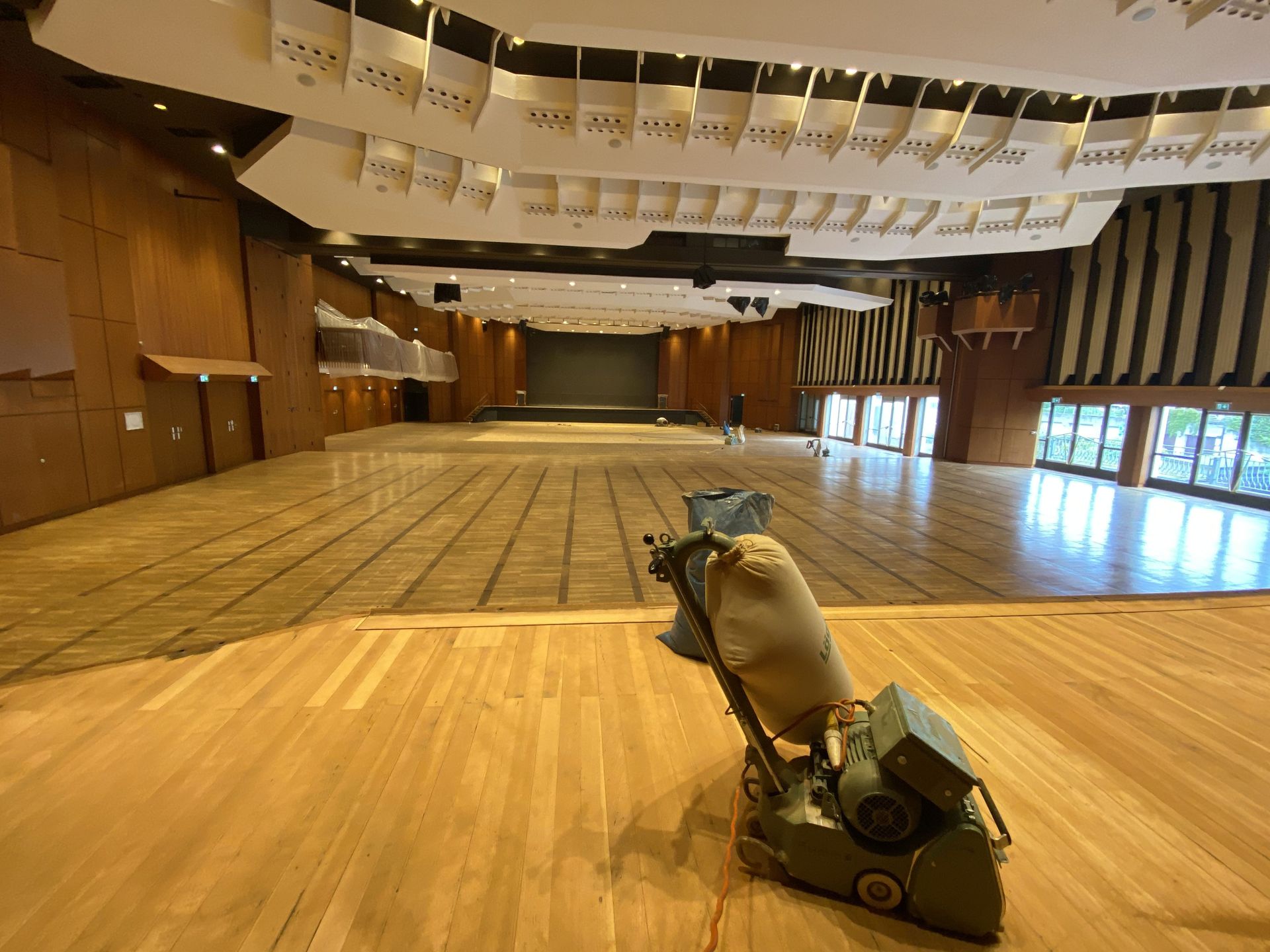 Bodenschleifmaschine in einem leeren Auditorium mit Holzvertäfelung, Fenstern und Bühne.