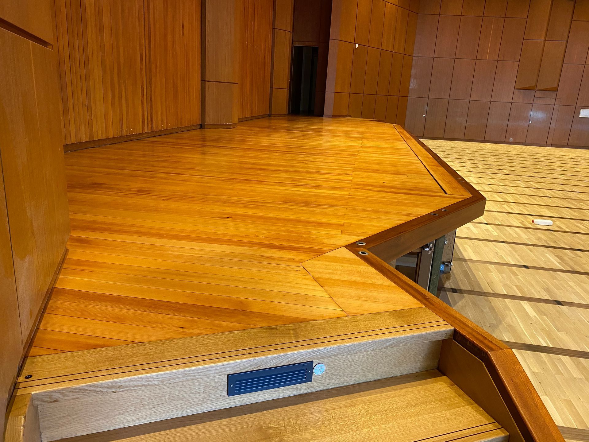 Holzbühne mit Treppe, Geländer und strukturierter Wand in einem Auditorium.