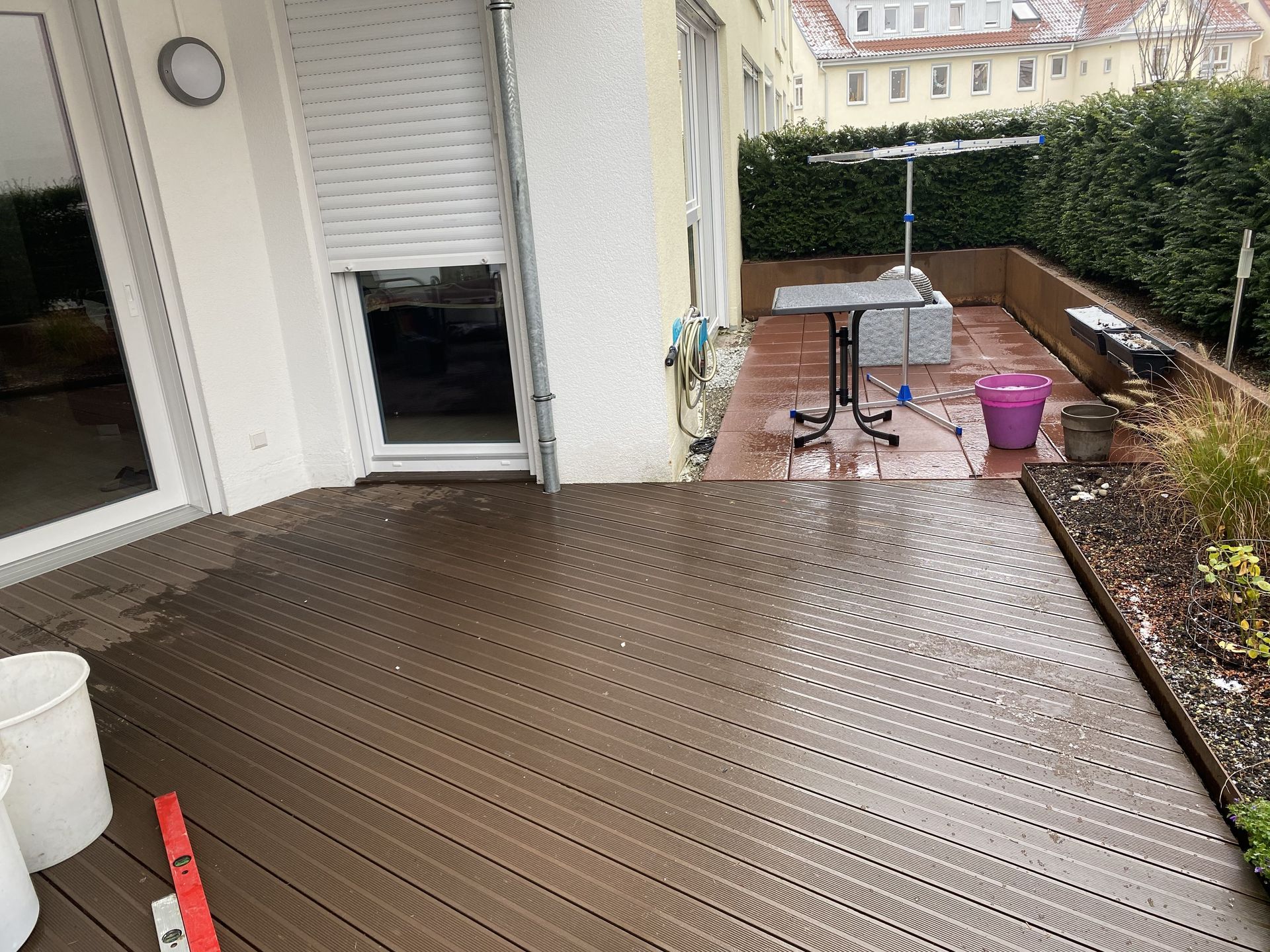 Ein braunes Verbunddeck auf einer Terrasse mit einem Tisch, Pflanzen und einem teilweise geschlossenen weißen Fensterladen.
