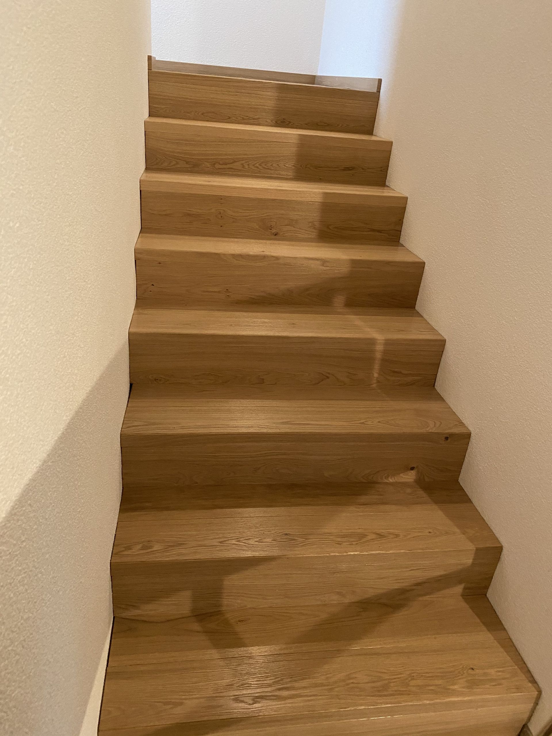 Holztreppe mit hellen Stufen und weißen Wänden. Blick von oben nach unten.