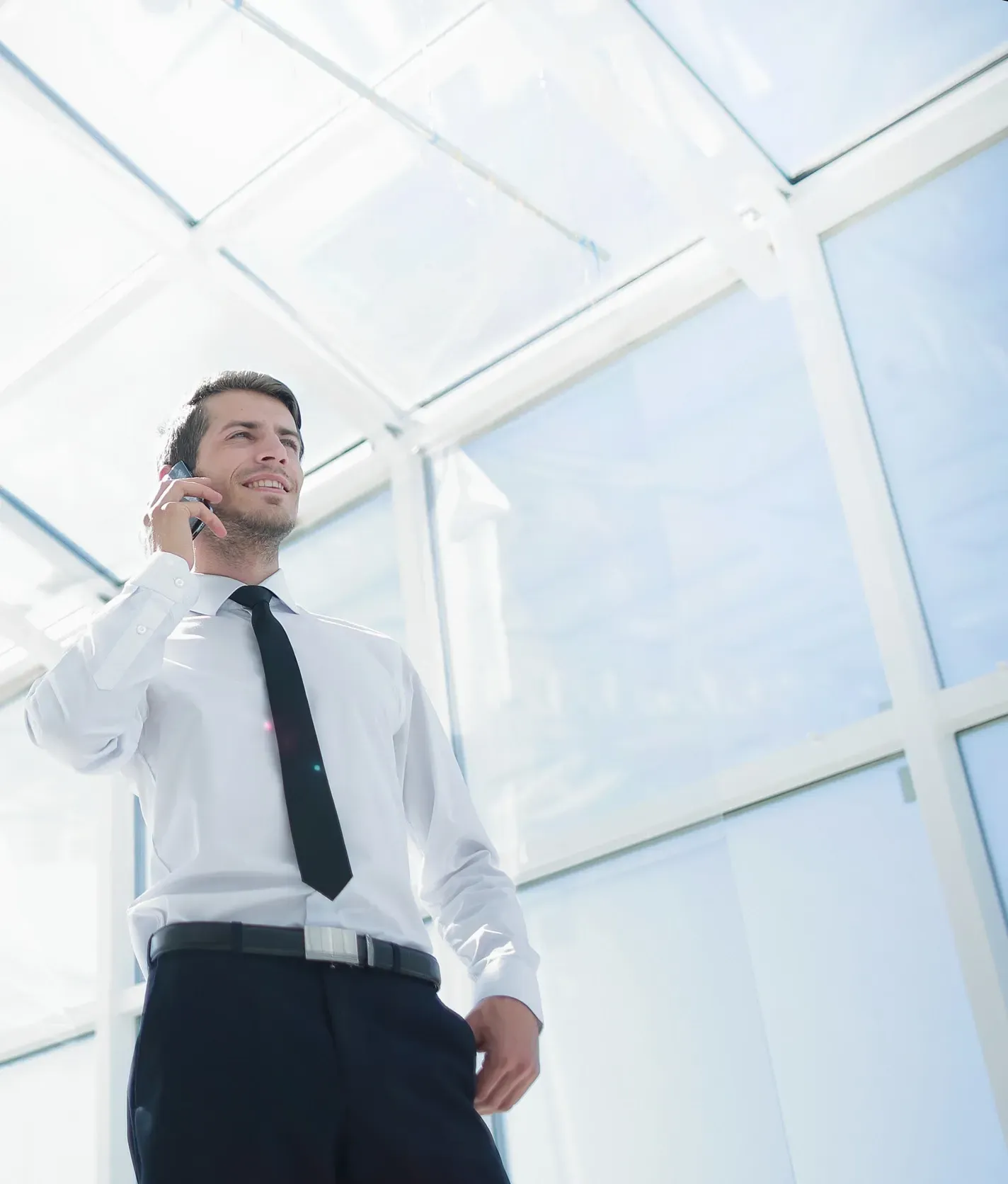 Hombre con camisa blanca y corbata negra hablando por teléfono, de pie frente a grandes ventanales.