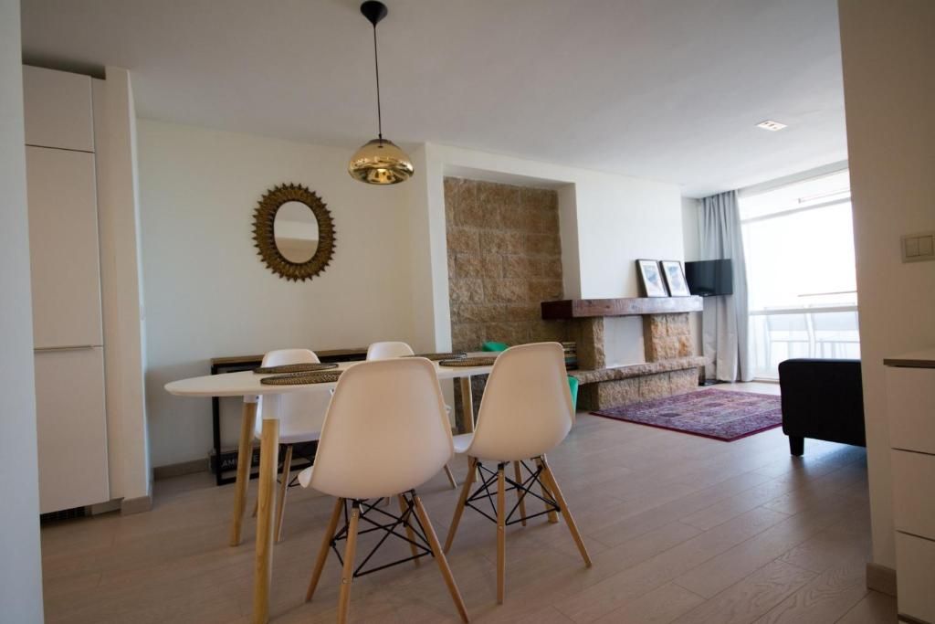 Comedor con mesa y sillas blancas, lámpara colgante dorada, espejo y chimenea de piedra.