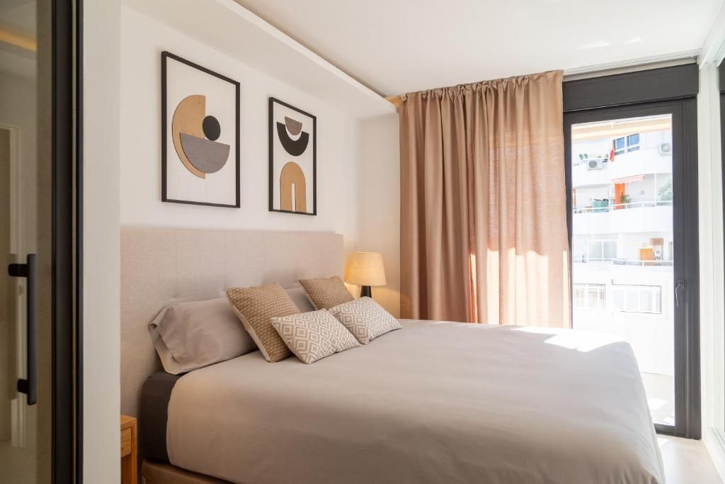 Interior de dormitorio con una cama, un espejo grande y un taburete pequeño.