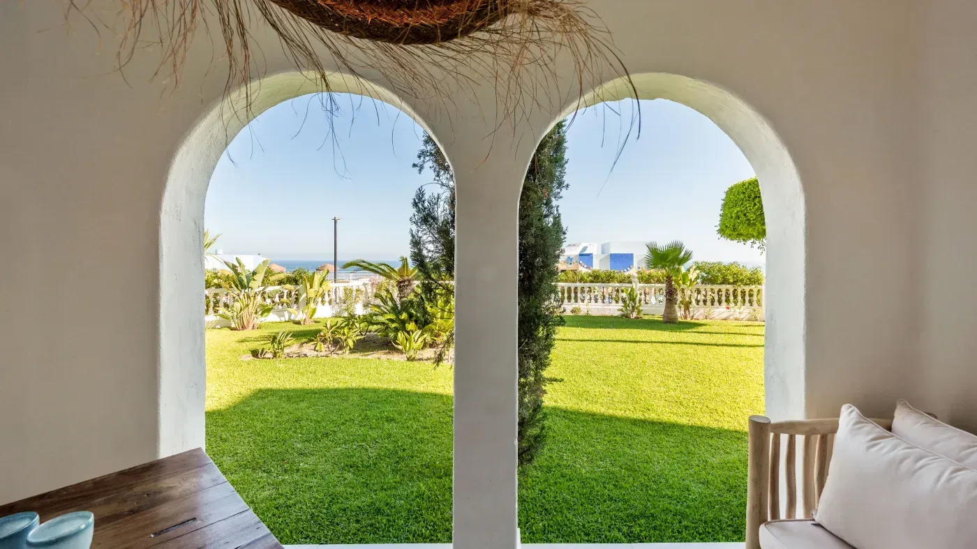 Dos ventanas arqueadas con vista a un césped verde y a edificios azules distantes bajo un cielo brillante.