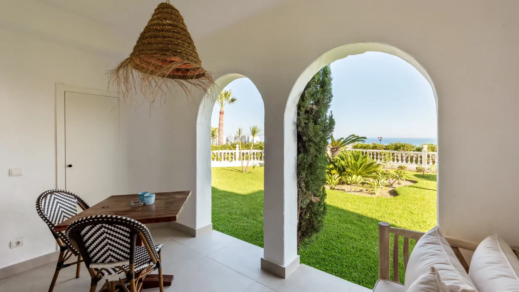 Un comedor con mesa y sillas, enmarcado por aberturas arqueadas con vista a un exuberante patio verde y al mar.