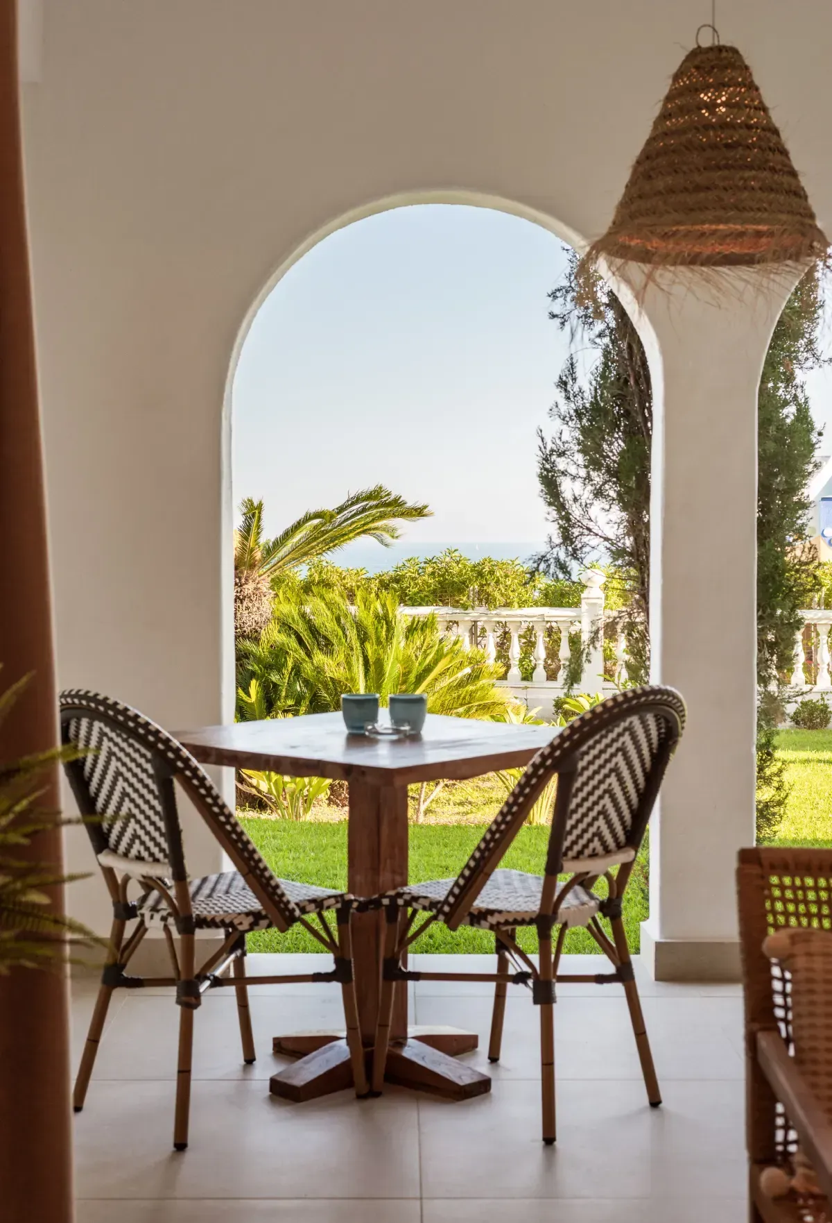 Mesa y sillas enmarcadas por una puerta arqueada con vista a un césped verde y al mar, con lámpara tejida.
