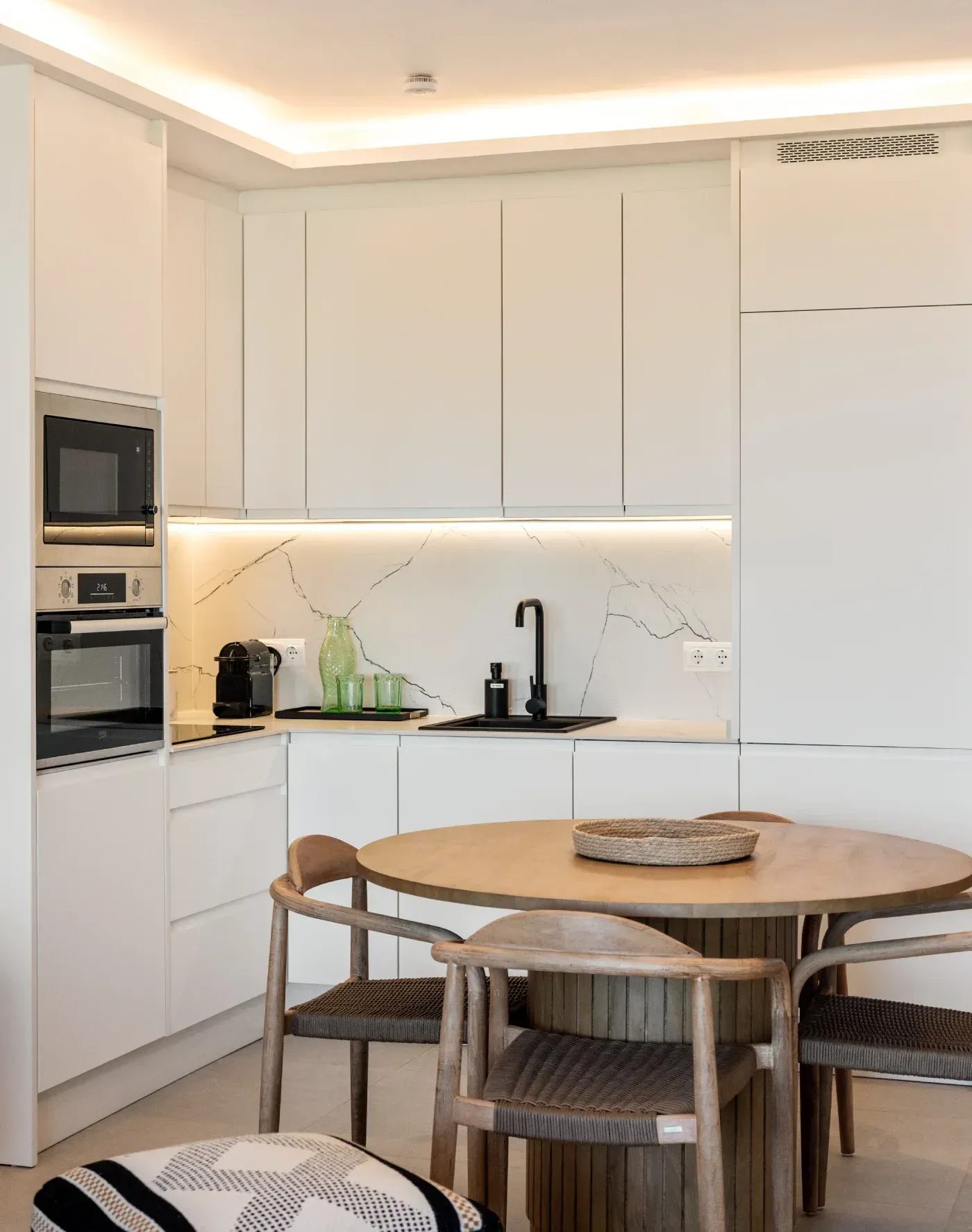 Cocina blanca moderna con detalles en negro, mesa redonda de madera, sillas y electrodomésticos empotrados.