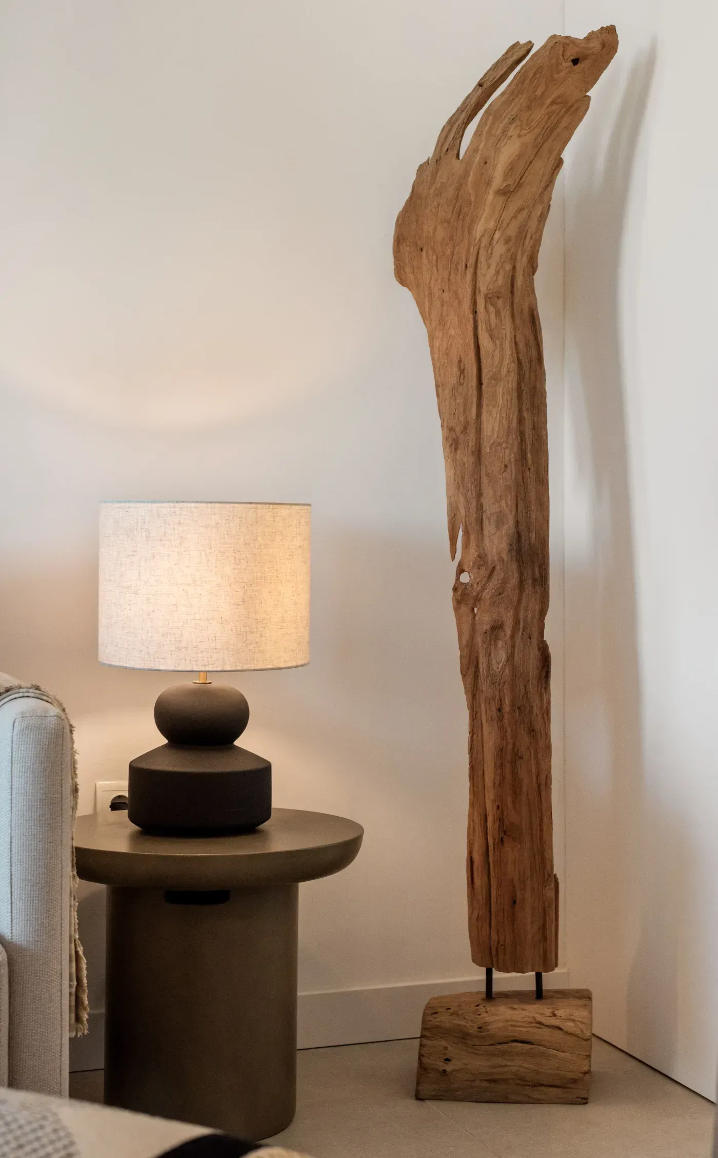 Lámpara sobre una mesa auxiliar junto a una escultura alta de madera desgastada. Ambiente en tonos neutros.