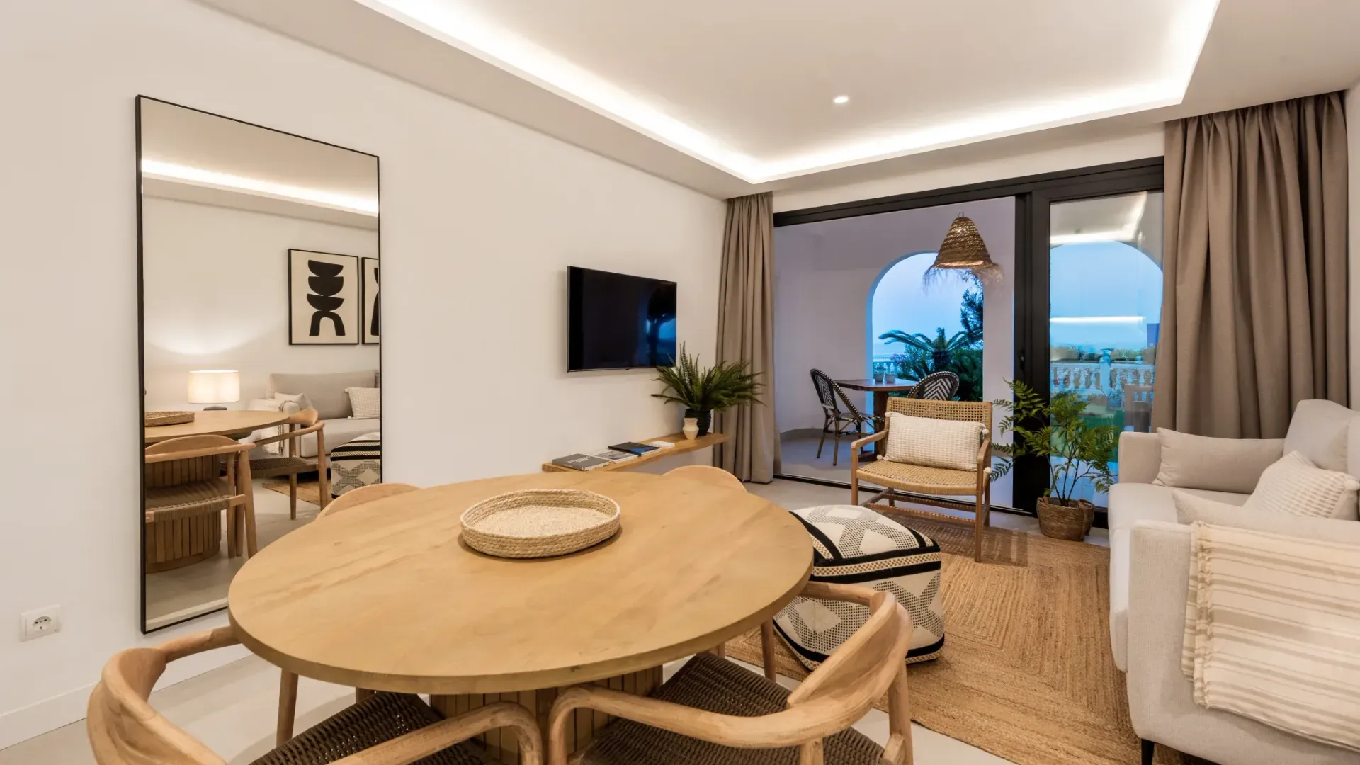 Sala de estar con mesa, sillas y sofá de madera. Hay un espejo de cuerpo entero y una puerta a un balcón.