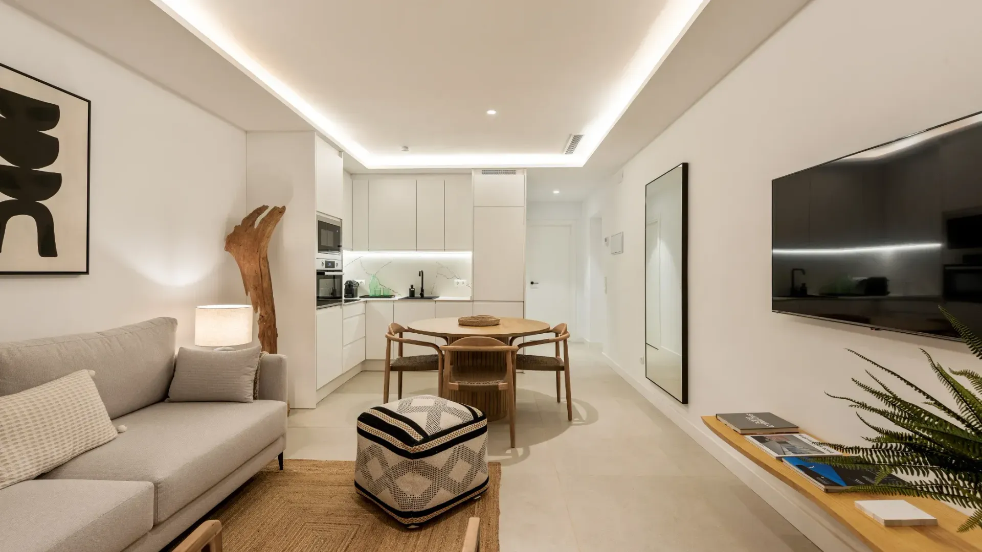 Espacio de estar moderno con sofá, mesa de comedor, cocina y televisión; paredes blancas y tonos neutros.