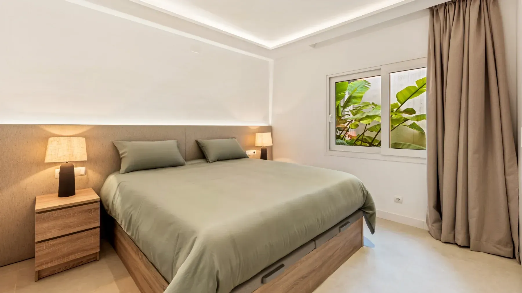 Dormitorio con cama, mesitas de noche, ventana y decoración neutra.