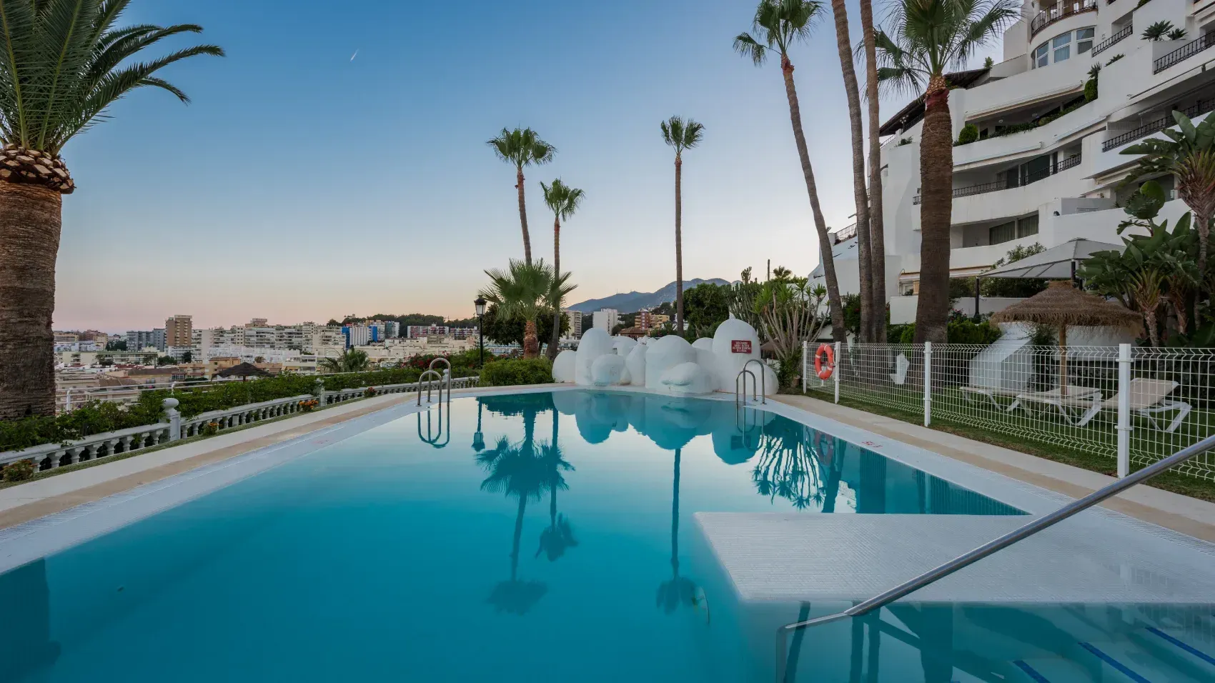 Piscina con agua cristalina, palmeras y edificios blancos al fondo.