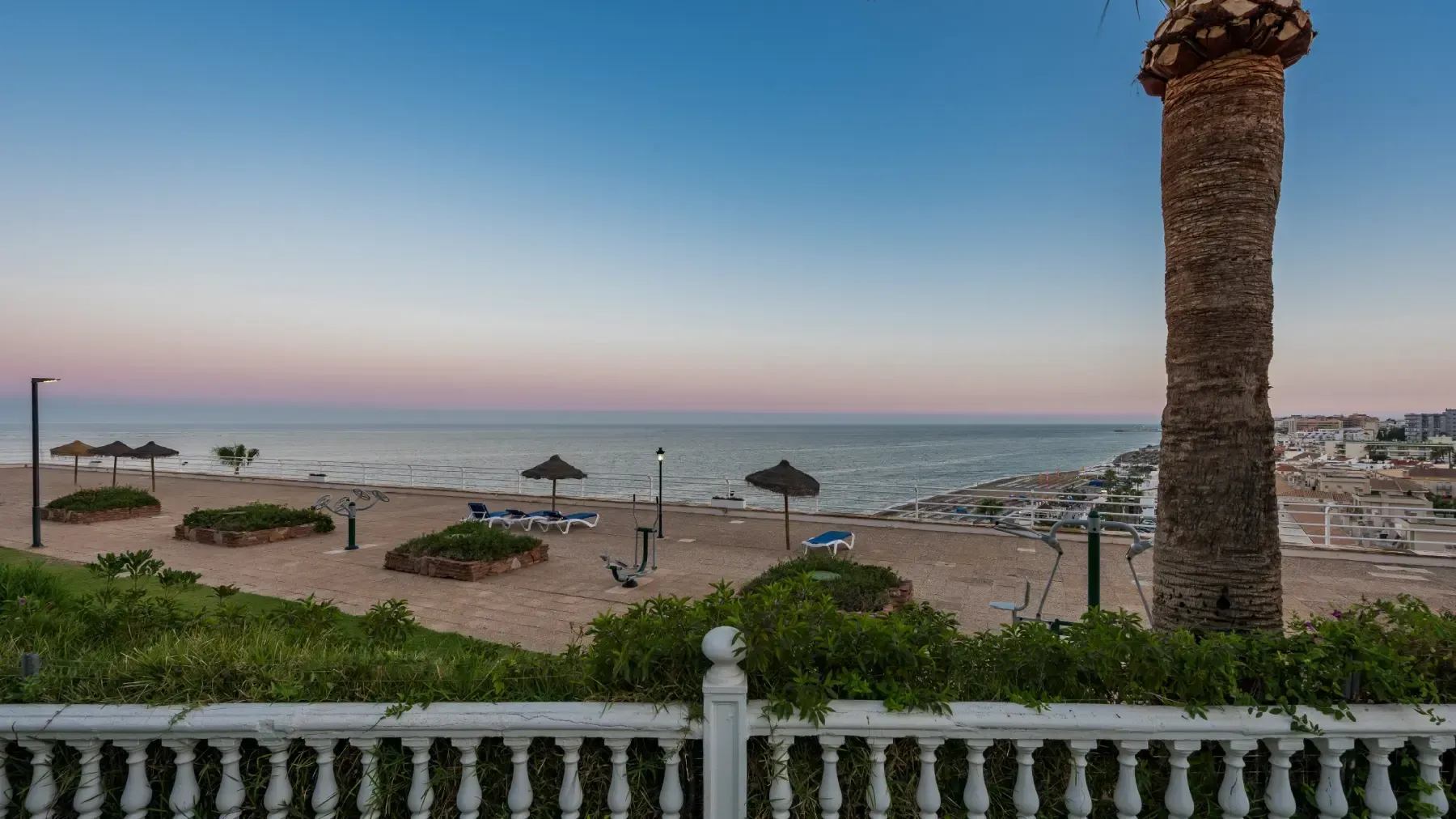 Vista de playa con arena, océano, palmeras y una cerca blanca bajo un cielo crepuscular.