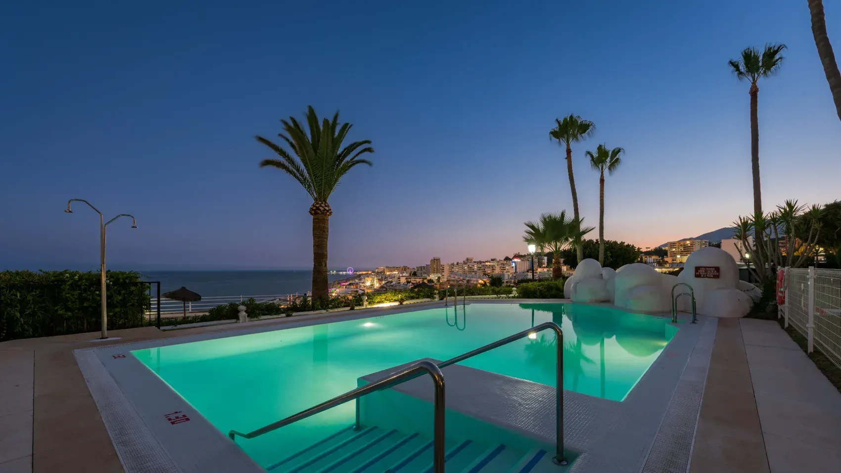 Piscina al atardecer con vistas a la playa y la ciudad. Palmeras rodean el agua bajo un cielo azul oscuro.