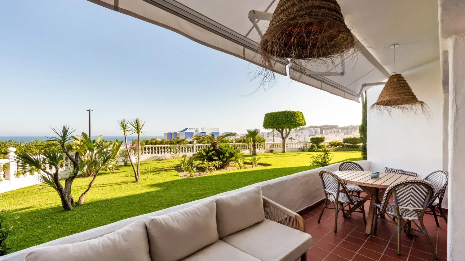 Balcón con sofá beige, mesa de madera y sillas de mimbre con vista a un césped verde y al mar.