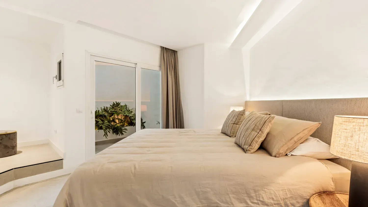 Dormitorio con cama grande, colores neutros, vista al balcón con naranjo.