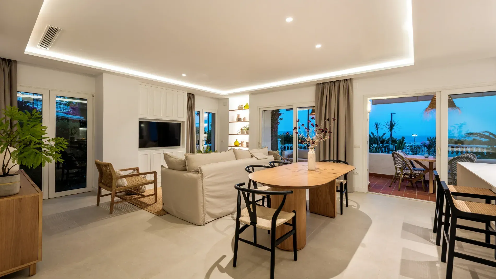 Luminosa sala de estar con paredes blancas, un sofá beige y una mesa de comedor de madera, con vista a un patio.