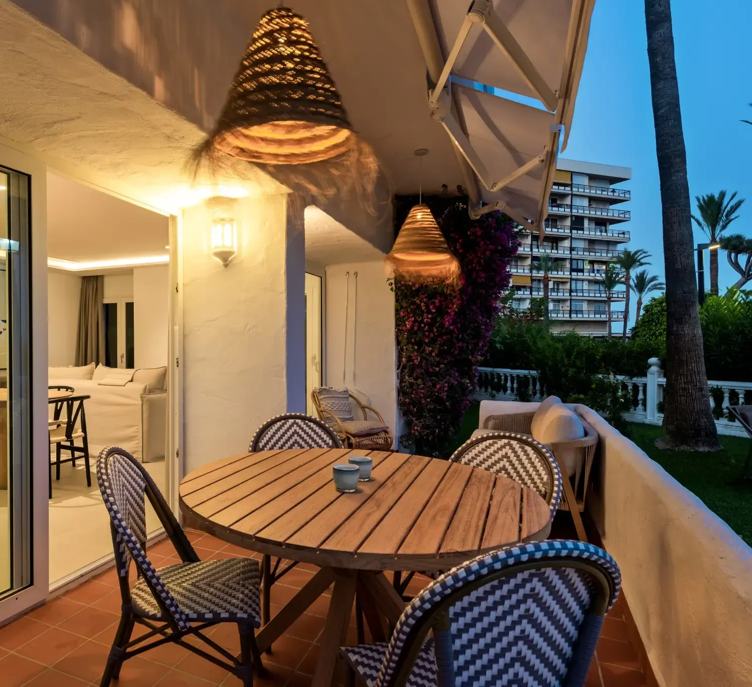 Patio exterior con mesa de madera, sillas y lámparas colgantes tejidas.