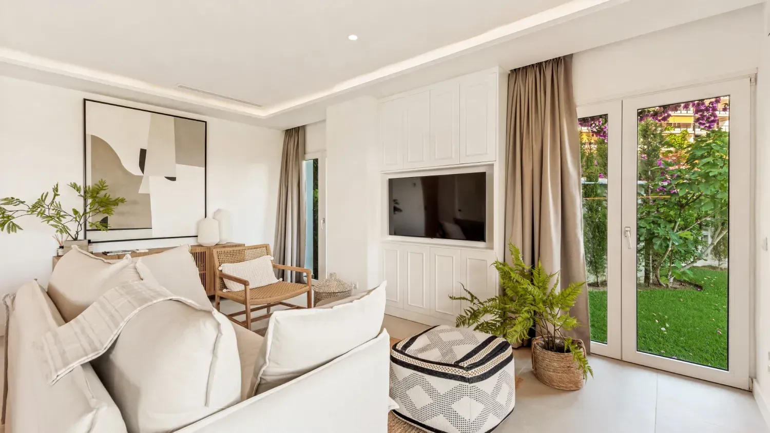 Sala de estar luminosa con paredes blancas, sofá color crema, TV y puertas francesas que conducen a un patio verde.