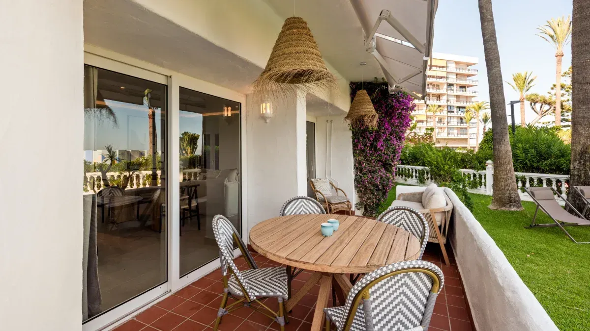 Balcón con mesa redonda de madera, sillas y pantalla de lámpara tejida. Vistas a la zona verde y al mar.