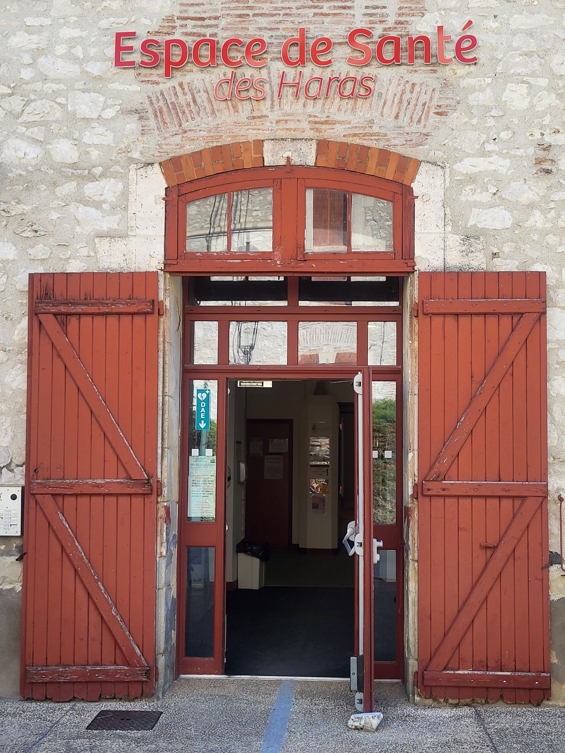 L'entrée en pierre de l'Espace de Santé des Haras avec des volets en bois rouge s'ouvre sur une porte intérieure.