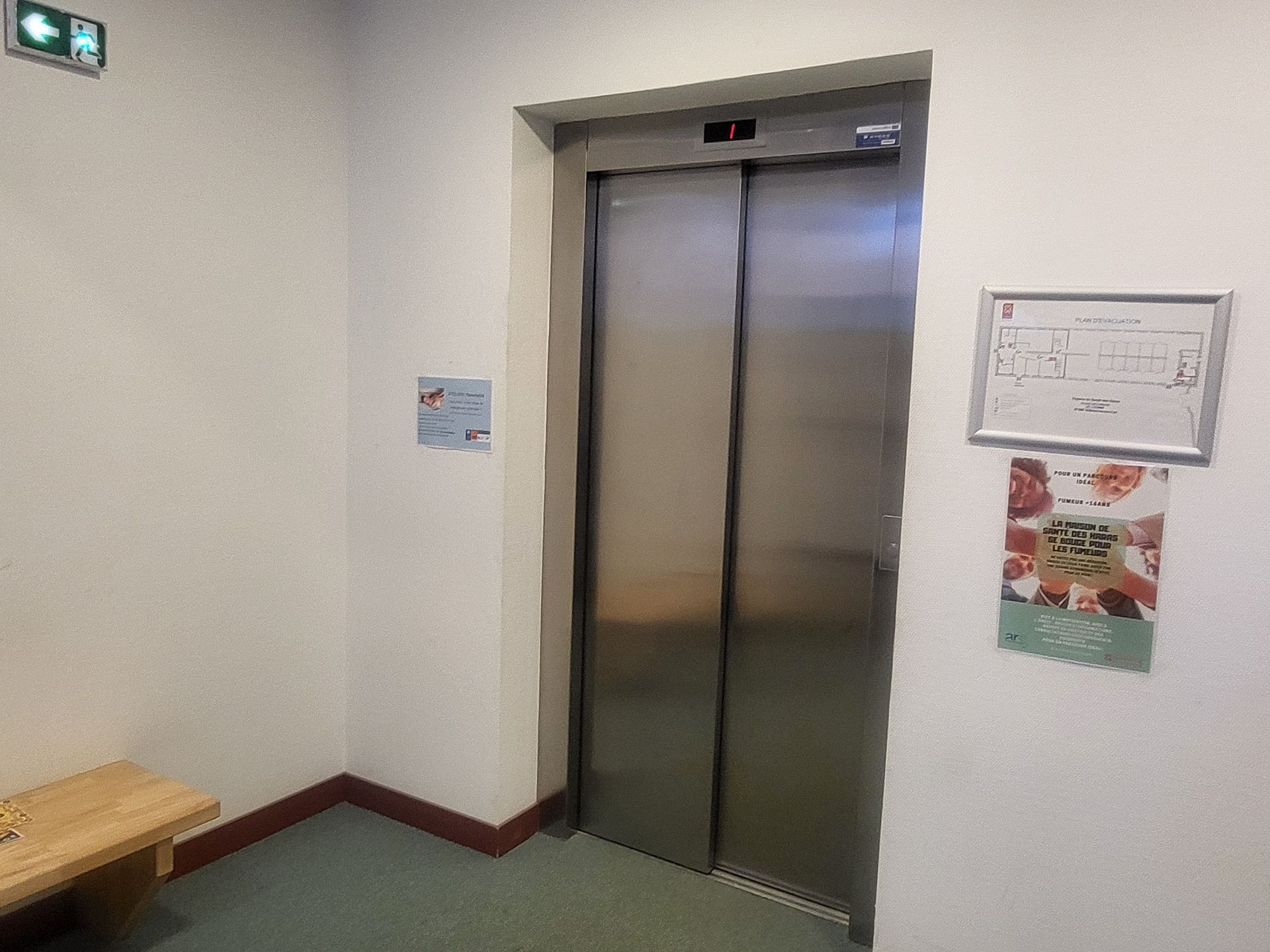 Un couloir avec un ascenseur métallique, un banc en bois clair, un petit panneau vert de sortie de secours et deux affiches murales encadrées.