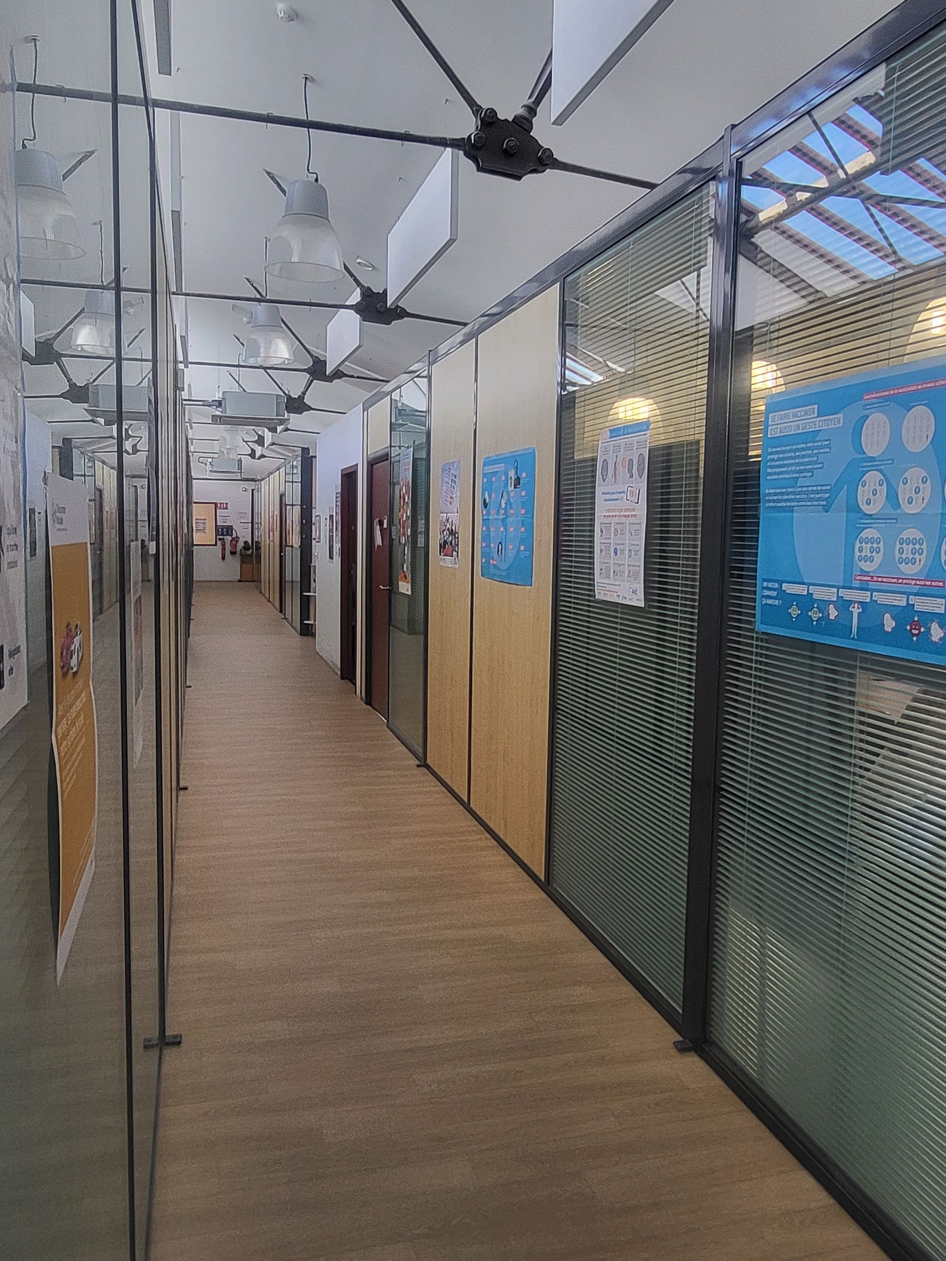 Un long couloir étroit au sol en bois, bordé de cloisons vitrées ornées d'affiches et de textures en maille.