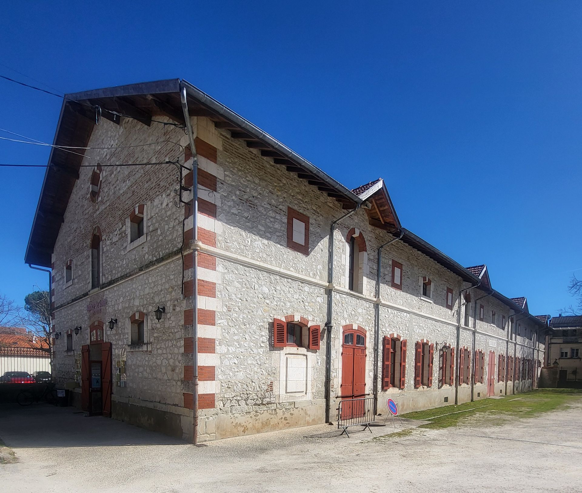 Une longue grange en pierre, aux angles et volets en briques rouges, se dresse sous un ciel d'un bleu éclatant par une journée claire.