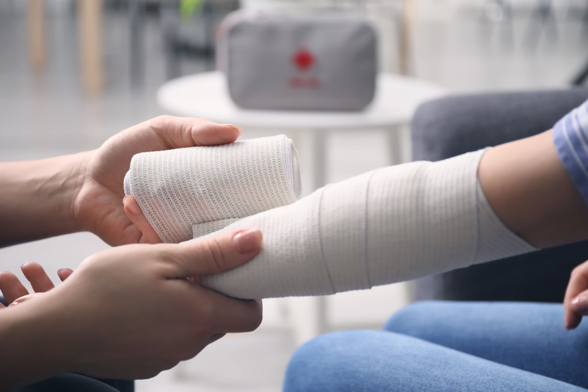 Les mains d'une personne enroulant un bandage élastique blanc autour de l'avant-bras d'une autre personne, avec une trousse de premiers secours en arrière-plan.