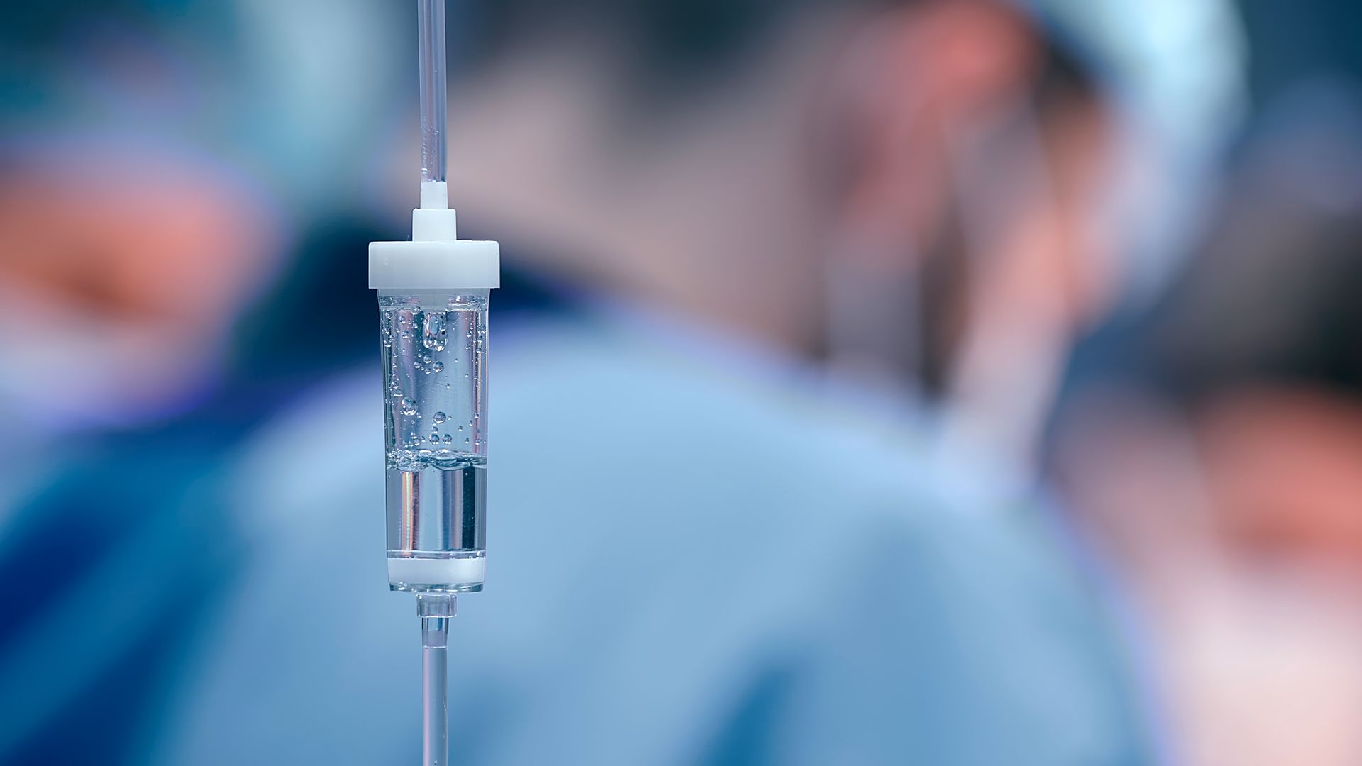 Gros plan sur une chambre de perfusion intraveineuse contenant un liquide transparent, sur fond d'environnement médical flou avec du personnel en blouse médicale.