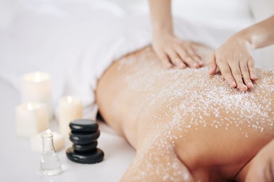 Persona recibiendo una exfoliación de espalda en un spa. Manos aplicándose el exfoliante, con velas cerca.
