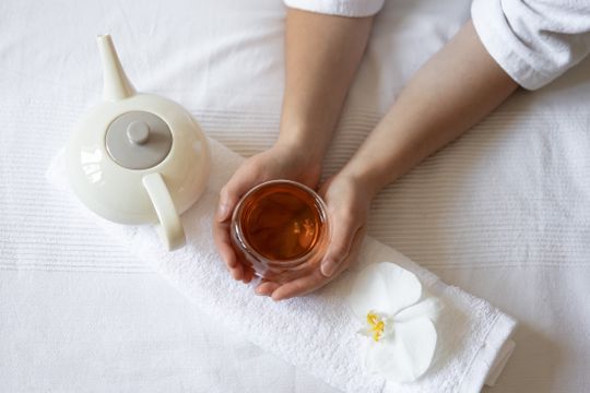 Manos sosteniendo un vaso de té sobre una toalla blanca, junto a una tetera y una flor de orquídea.