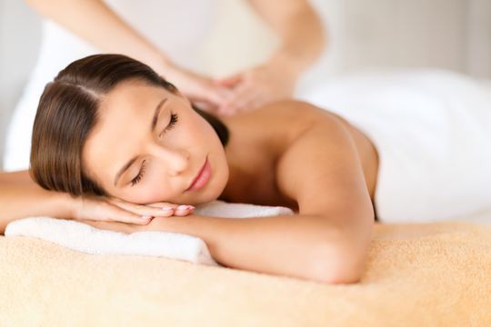 Mujer recibiendo un masaje en un spa; ojos cerrados, manos relajadas, siendo masajeada en la espalda.